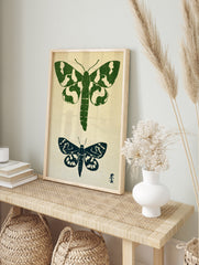 Two moth illustrations in green and navy block-print style on a textured neutral background, in an alternate framed mockup
