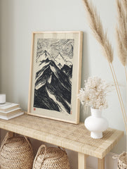 Black and white woodcut style print of a dramatic mountain range with swirling, textured sky, in an alternate framed mockup