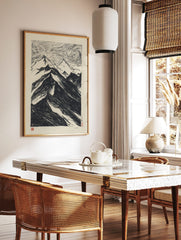 Black and white woodcut style print of a dramatic mountain range with swirling, textured sky, in a styled room mockup