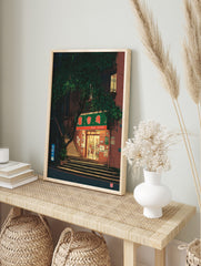 Illustrative print of a shop entrance at night, with a green sign, steps, and a large tree, in an alternate framed mockup