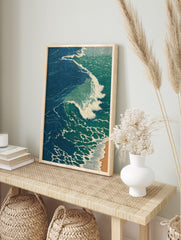 Illustrative print of a large ocean wave with blue-green water and white foam breaking onto a, in an alternate framed mockup