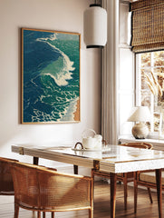 Illustrative print of a large ocean wave with blue-green water and white foam breaking onto a sandy, in a styled room mockup