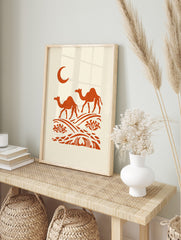 Illustrative orange dromedary camels walking on desert dunes under a crescent moon on a cream, in an alternate framed mockup