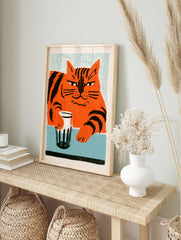 Stylised orange cat with black stripes holding a glass on a blue table, illustrative wall art, in an alternate framed mockup
