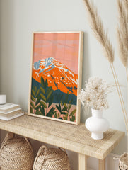 Illustrative orange mountain with white snow, pink sky, and dark green foliage in foreground, in an alternate framed mockup