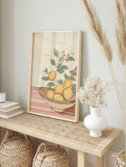 Illustrative orange still life print with fruit in a bowl, green leaves, and red berries on a, in an alternate framed mockup