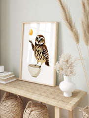 Illustrated wall art print of a brown speckled owl balancing a golden egg above a white bowl, in an alternate framed mockup