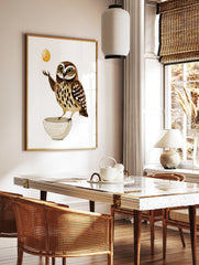 Illustrated wall art print of a brown speckled owl balancing a golden egg above a white bowl on a, in a styled room mockup