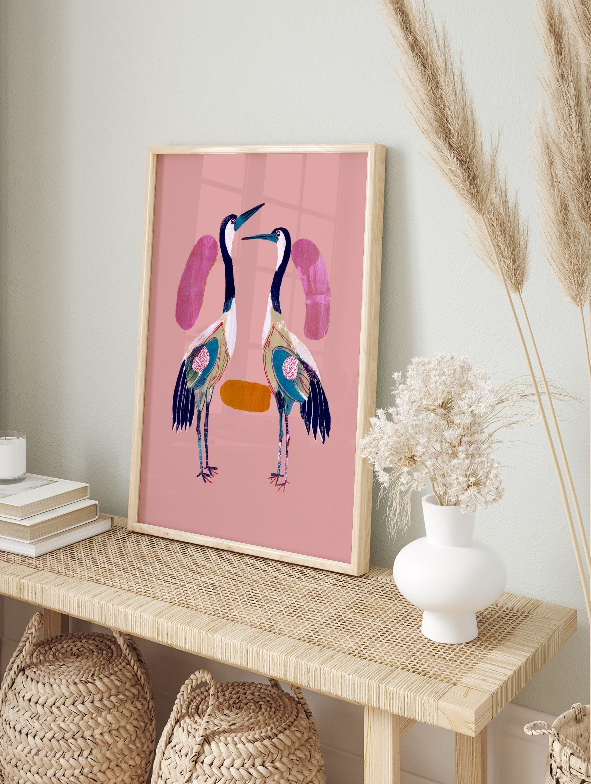 Two stylised cranes with navy, white, teal plumage on a pink background, facing each other, in an alternate framed mockup