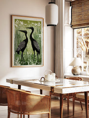 Linocut style art print of two black cranes standing in green reeds with white flowers and water, in a styled room mockup