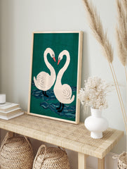 Two white swans with pink beaks form a heart shape against a teal-green background with blue, in an alternate framed mockup