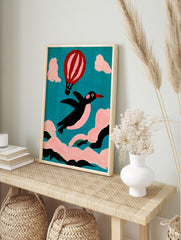Whimsical illustration of a black penguin flying with a pink hot air balloon against a teal, in an alternate framed mockup
