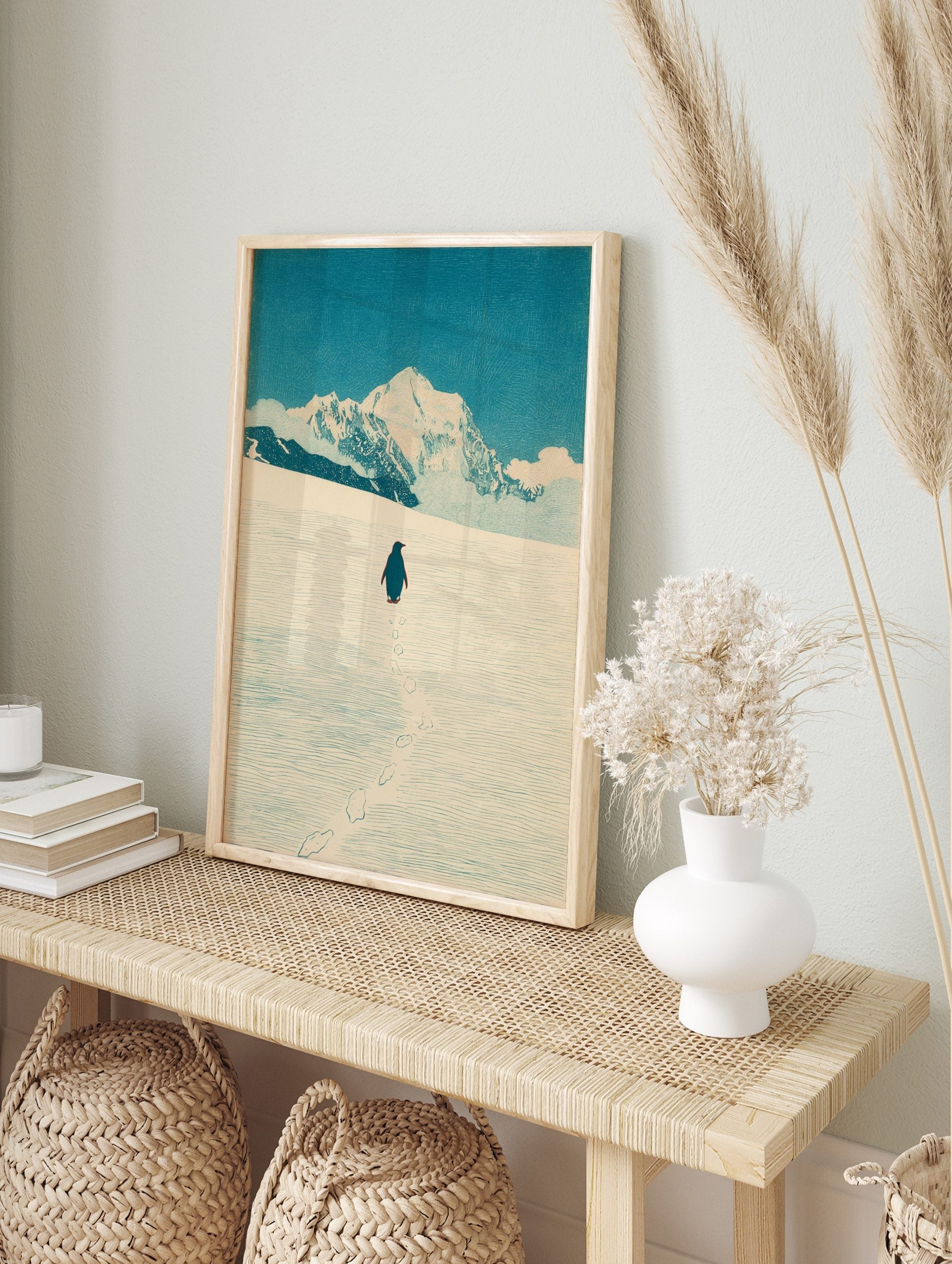 Illustrative print of a penguin walking in snow towards large, white mountains under a blue, in an alternate framed mockup