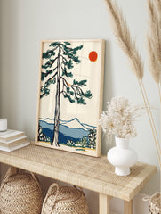 Illustrative pine tree with dark green foliage, red sun, blue mountains, and water in a, in an alternate framed mockup