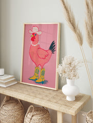 Pink chicken wearing a white cowboy hat, pearl necklace, and yellow cowboy boots on a pink, in an alternate framed mockup