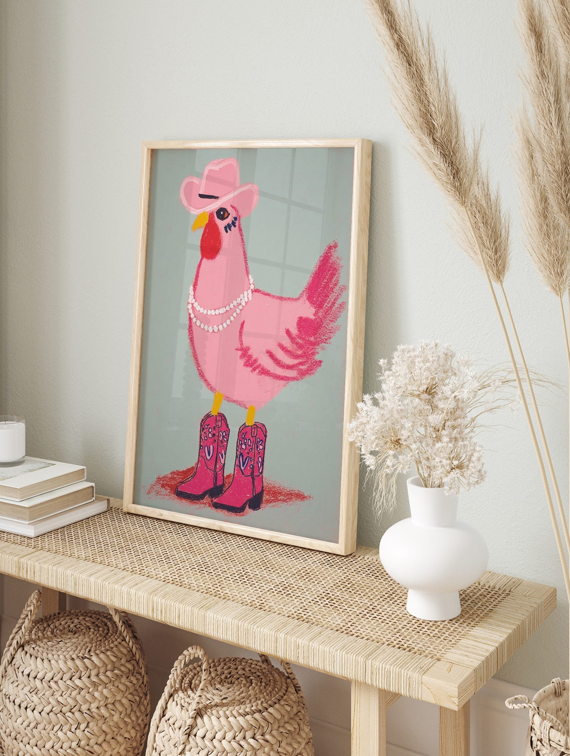 Illustrative pink chicken wearing a cowboy hat, pearl necklace, and pink cowboy boots on a, in an alternate framed mockup