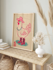 Illustrative print of a pink cowgirl chicken wearing a cowboy hat, beaded necklace, and boots, in an alternate framed mockup