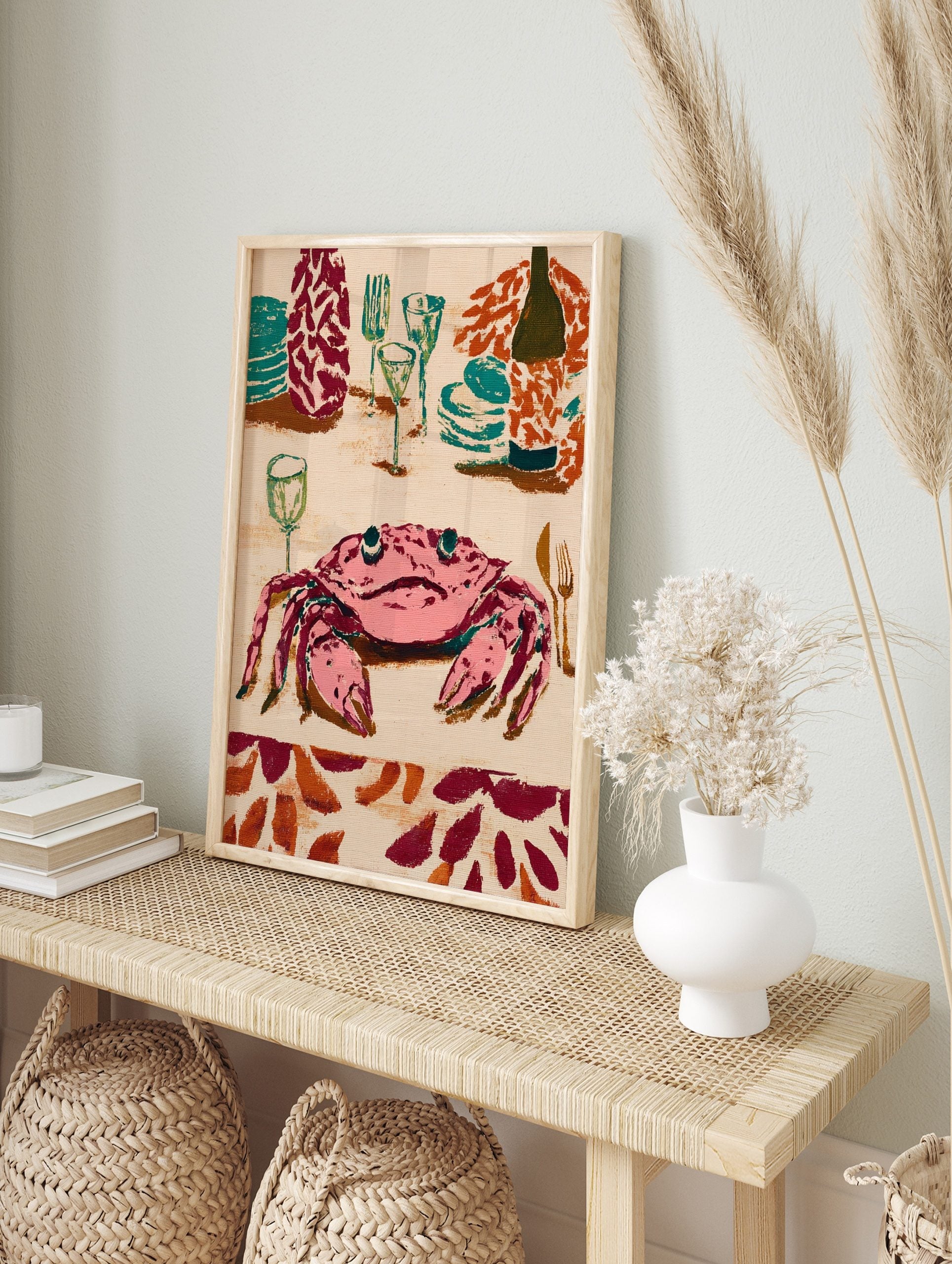 Illustrative wall art print of a pink crab at a dinner table with teal plates, glasses, and a, in an alternate framed mockup