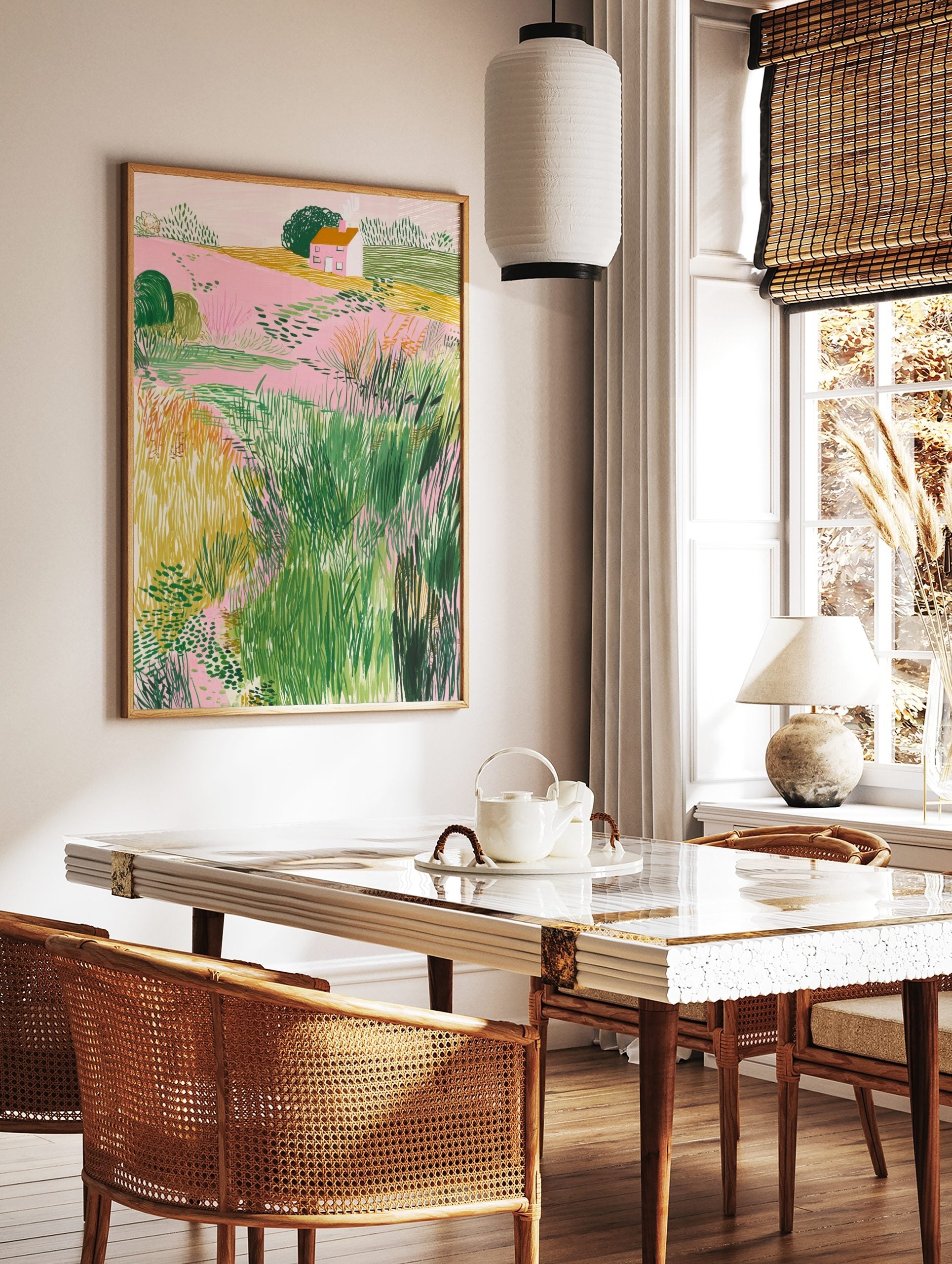 Illustrative landscape art print with pink fields, green bushes, yellow grass, and a small house, in a styled room mockup