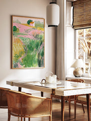 Illustrative landscape art print with pink fields, green bushes, yellow grass, and a small house, in a styled room mockup