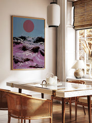 Surreal art print of pink ocean waves crashing with white foam under a large red sun in a blue sky, in a styled room mockup