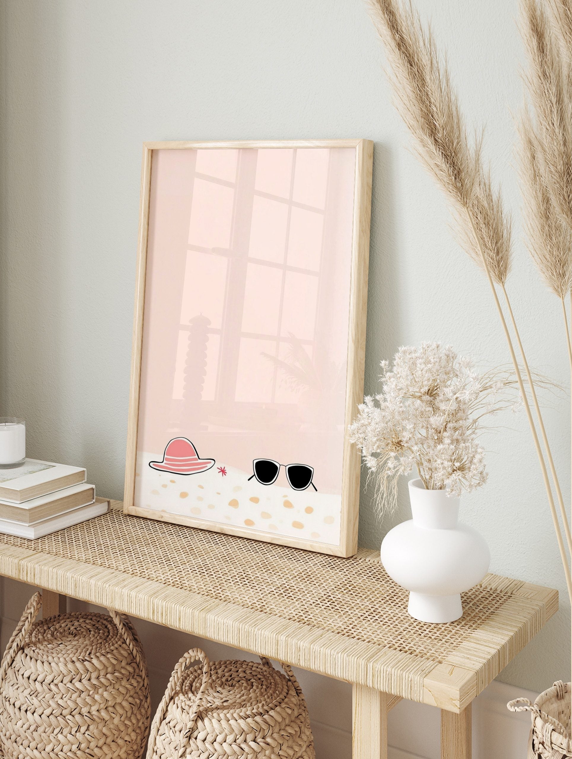 Illustrative print of a pink sun hat, black sunglasses, and red starfish on sand with a peach, in an alternate framed mockup