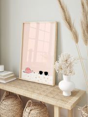 Illustrative print of a pink sun hat, black sunglasses, and red starfish on sand with a peach, in an alternate framed mockup