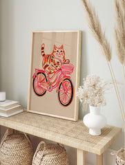 Illustrative print of an orange tabby cat riding a pink and red bicycle with a striped basket, in an alternate framed mockup