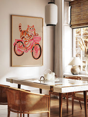 Illustrative print of an orange tabby cat riding a pink and red bicycle with a striped basket, in a styled room mockup