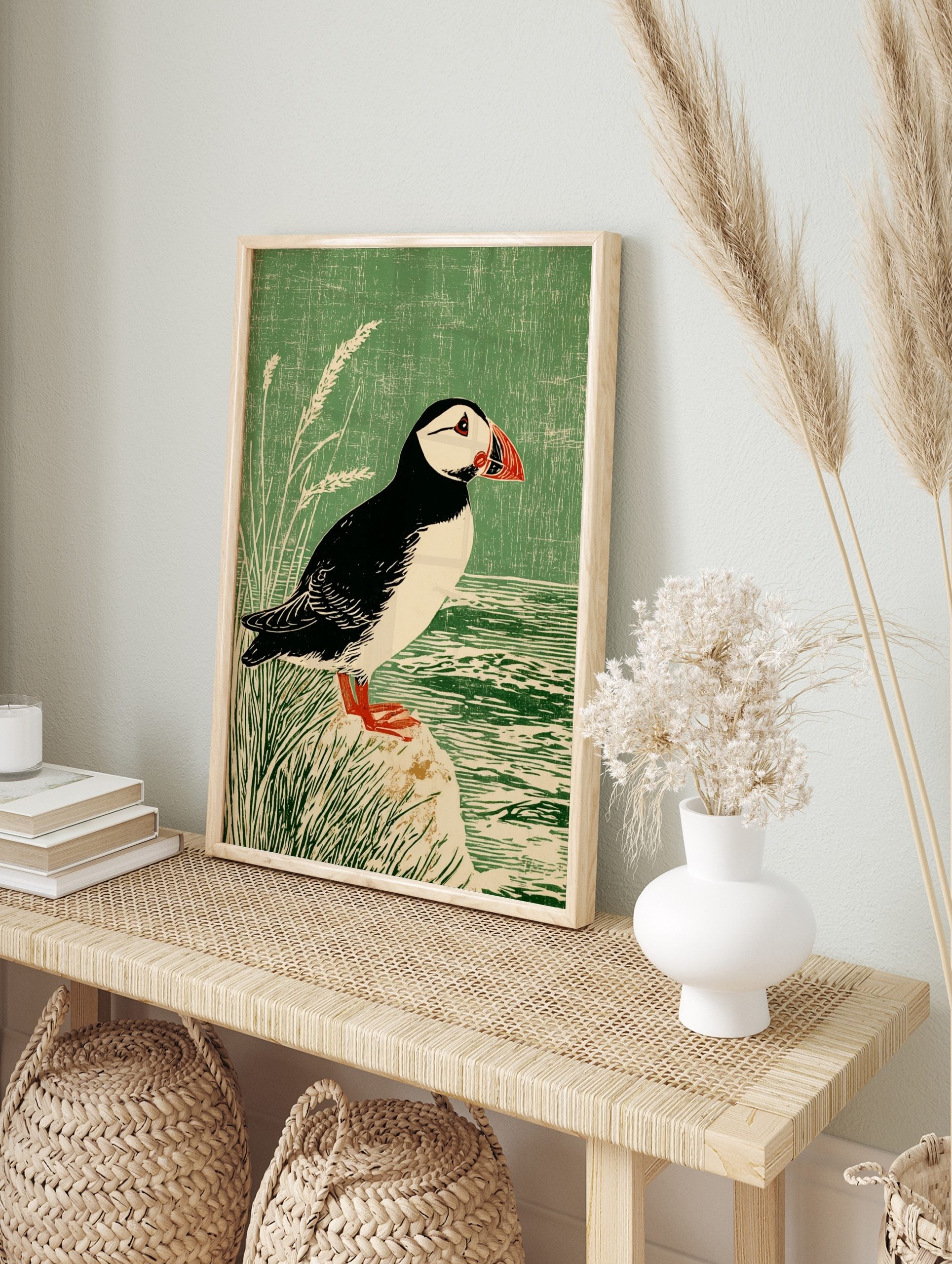 Illustrative print of a puffin bird with a red beak, standing on a rock by water and green, in an alternate framed mockup