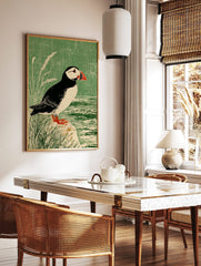 Illustrative print of a puffin bird with a red beak, standing on a rock by water and green grass, in a styled room mockup