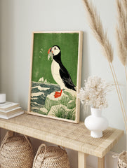 Illustrative woodcut style print of a puffin holding fish in its beak, standing on a rock by, in an alternate framed mockup