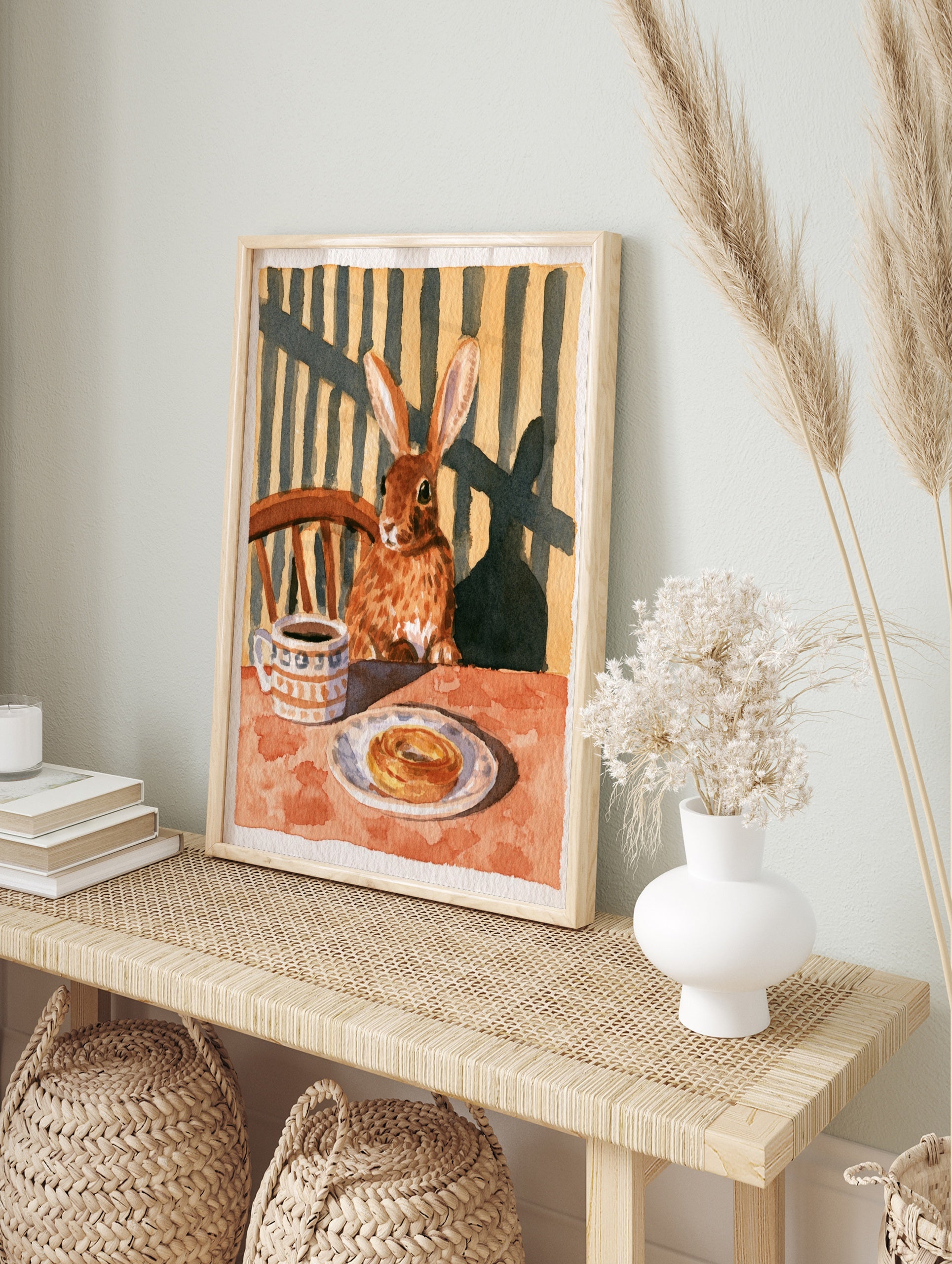 Illustration of a brown rabbit sitting at a table with coffee and a pastry, warm earthy, in an alternate framed mockup