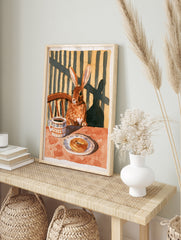 Illustration of a brown rabbit sitting at a table with coffee and a pastry, warm earthy, in an alternate framed mockup