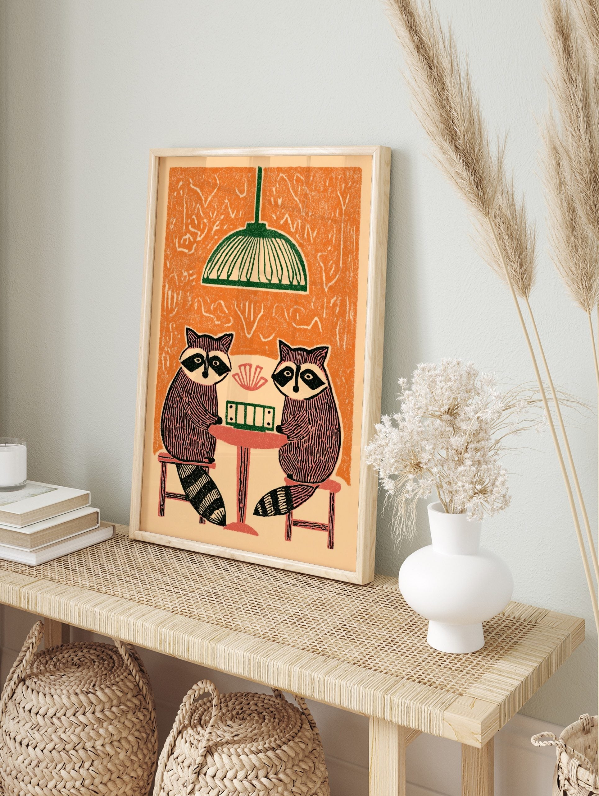 Illustrative print of two raccoons playing cards at a table under a green lamp, with an, in an alternate framed mockup