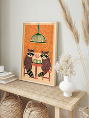 Illustrative print of two raccoons playing cards at a table under a green lamp, with an, in an alternate framed mockup