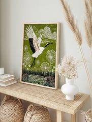 Red-crowned crane in flight over green landscape with white dandelion seed heads and water, in an alternate framed mockup