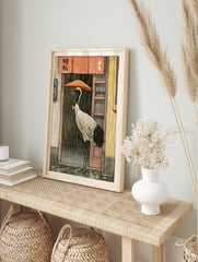 Red-crowned crane holding an orange umbrella standing in the rain before a shop with a red, in an alternate framed mockup