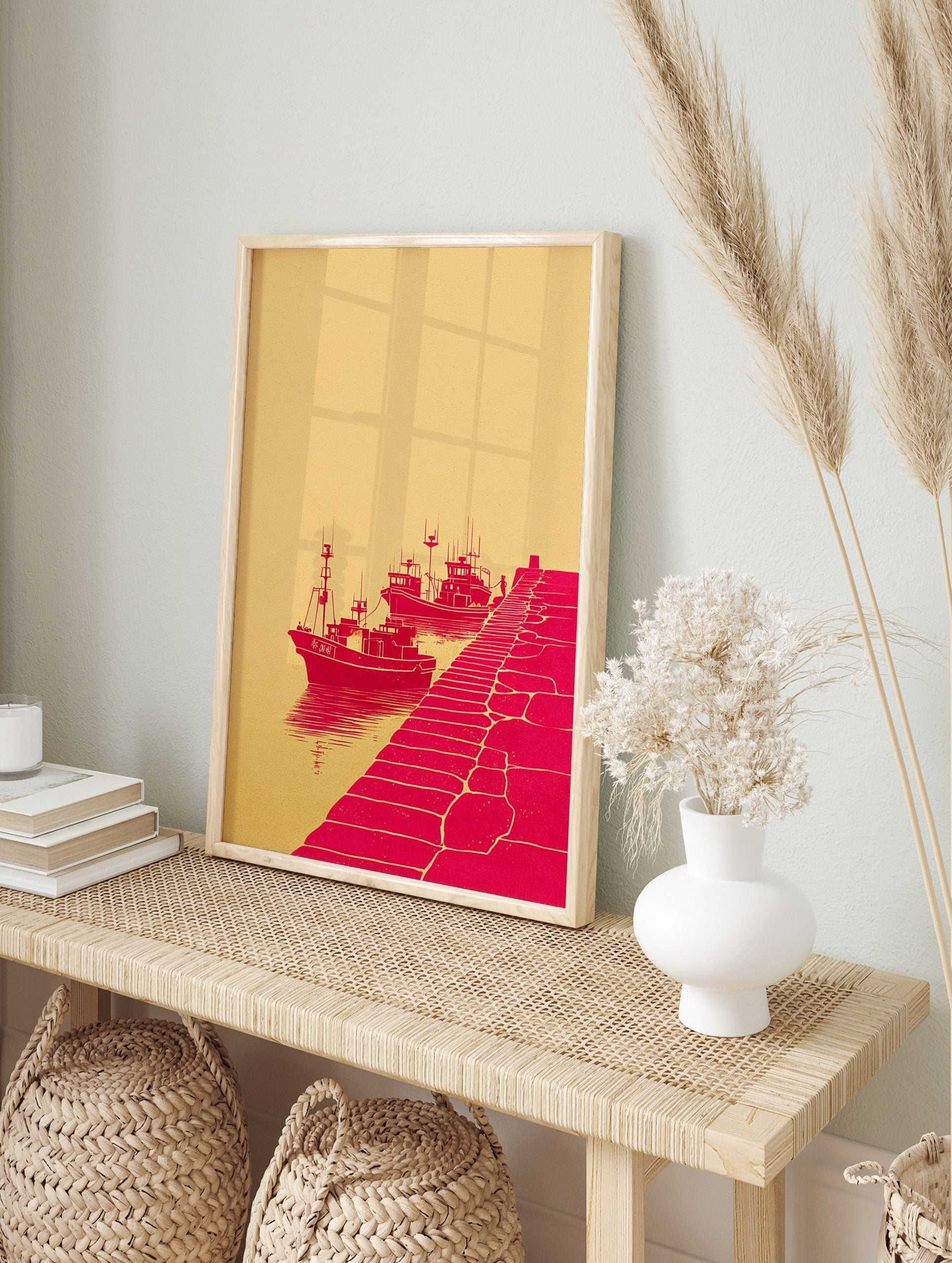Red illustrative print of three fishing boats docked by a pier with a figure, against a beige, in an alternate framed mockup