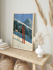 Red skis and poles in deep snow with a mountain cabin and forest background illustration, in an alternate framed mockup