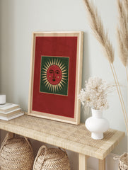 Stylized red sun face illustration with green background and cream rays, folk art style wall, in an alternate framed mockup