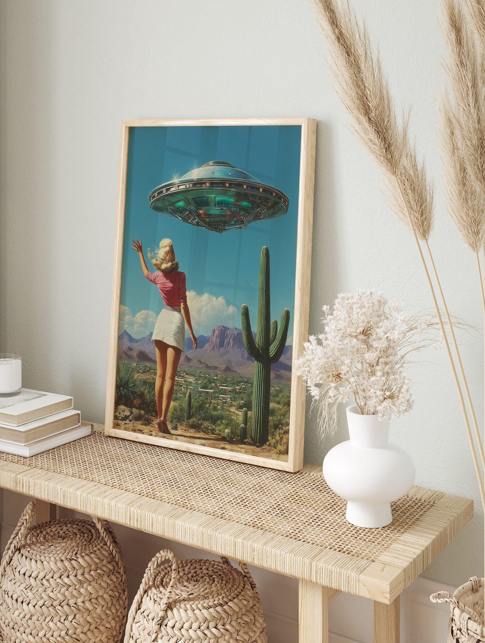 Retro UFO desert wall art featuring a woman waving at a flying saucer above a landscape with cacti and mountains, displayed as the same artwork in an alternate framed wall art mockup