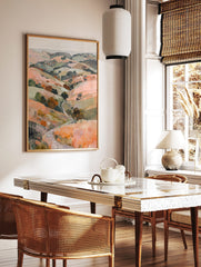 Painterly landscape print of rolling hills with a winding path and trees in peach, green, and, in a styled room mockup