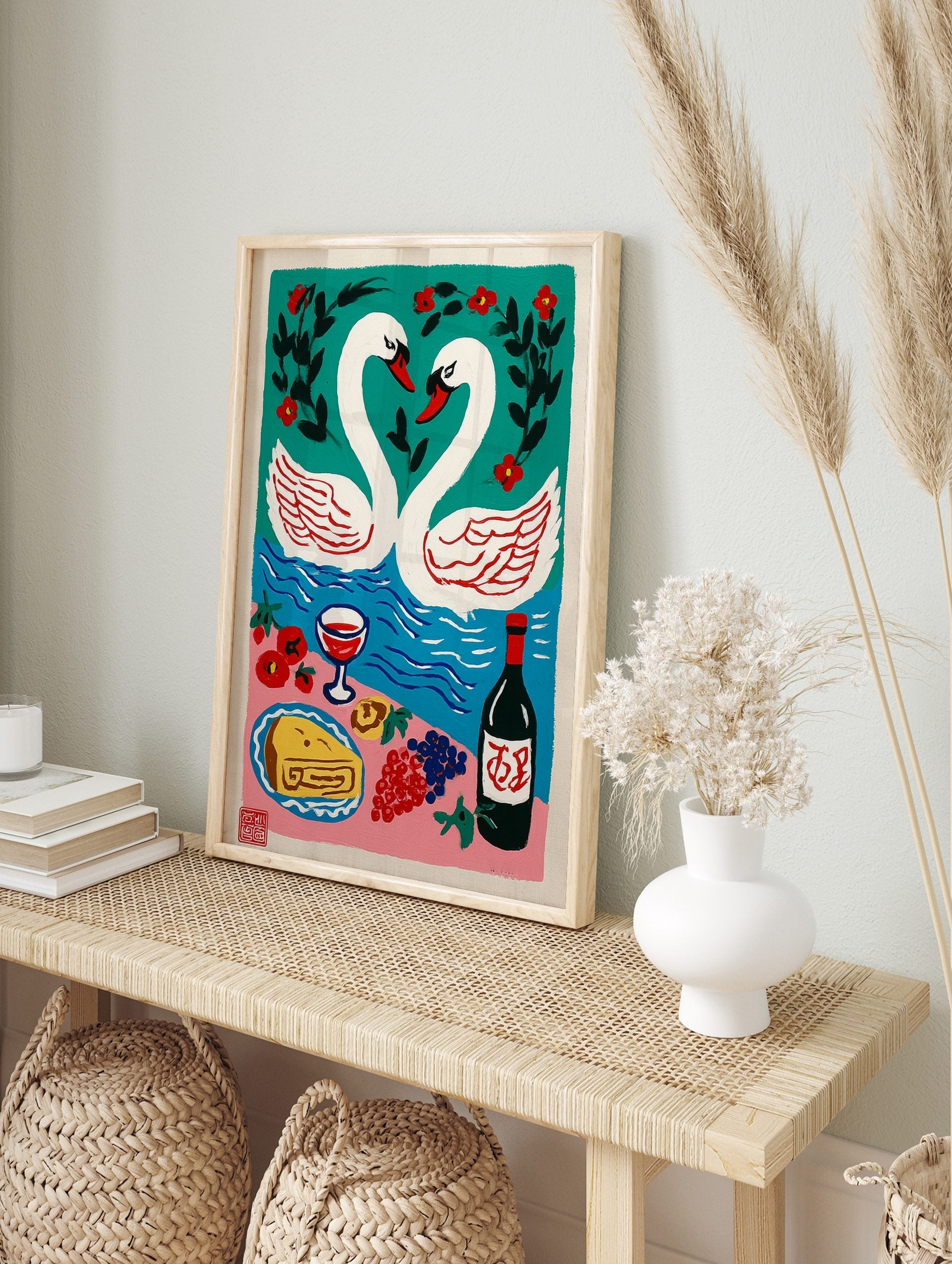 Illustrative print of two white swans forming a heart on teal water, with a picnic spread of, in an alternate framed mockup