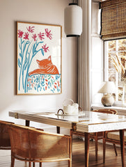 Illustrative print of a sleeping orange cat with red flowers and blue stems on a patterned surface, in a styled room mockup