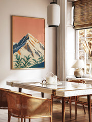 Illustrative snow-capped mountain print with peach sky and green foreground plants, in a styled room mockup