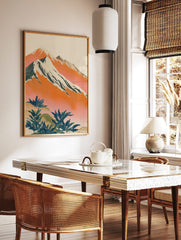 Illustrative mountain landscape art print with snow-capped peaks, orange and pink sky, and green, in a styled room mockup