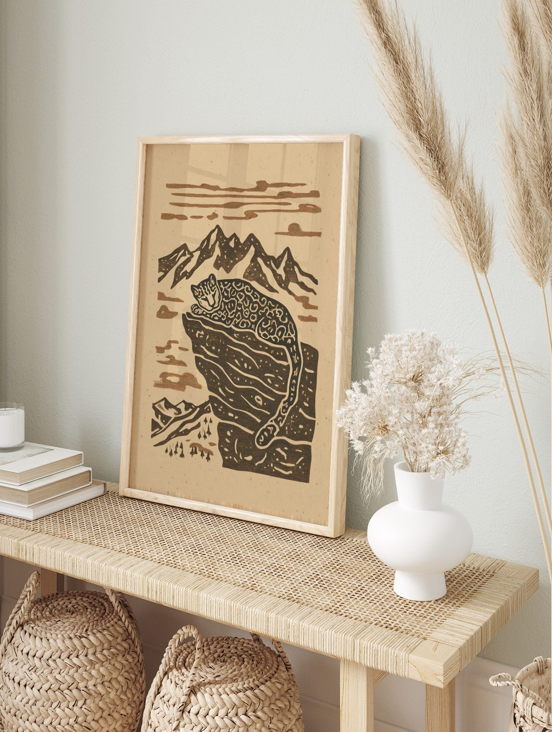 Stylised snow leopard resting on a rocky mountain ledge with peaks and clouds in a two-tone, in an alternate framed mockup