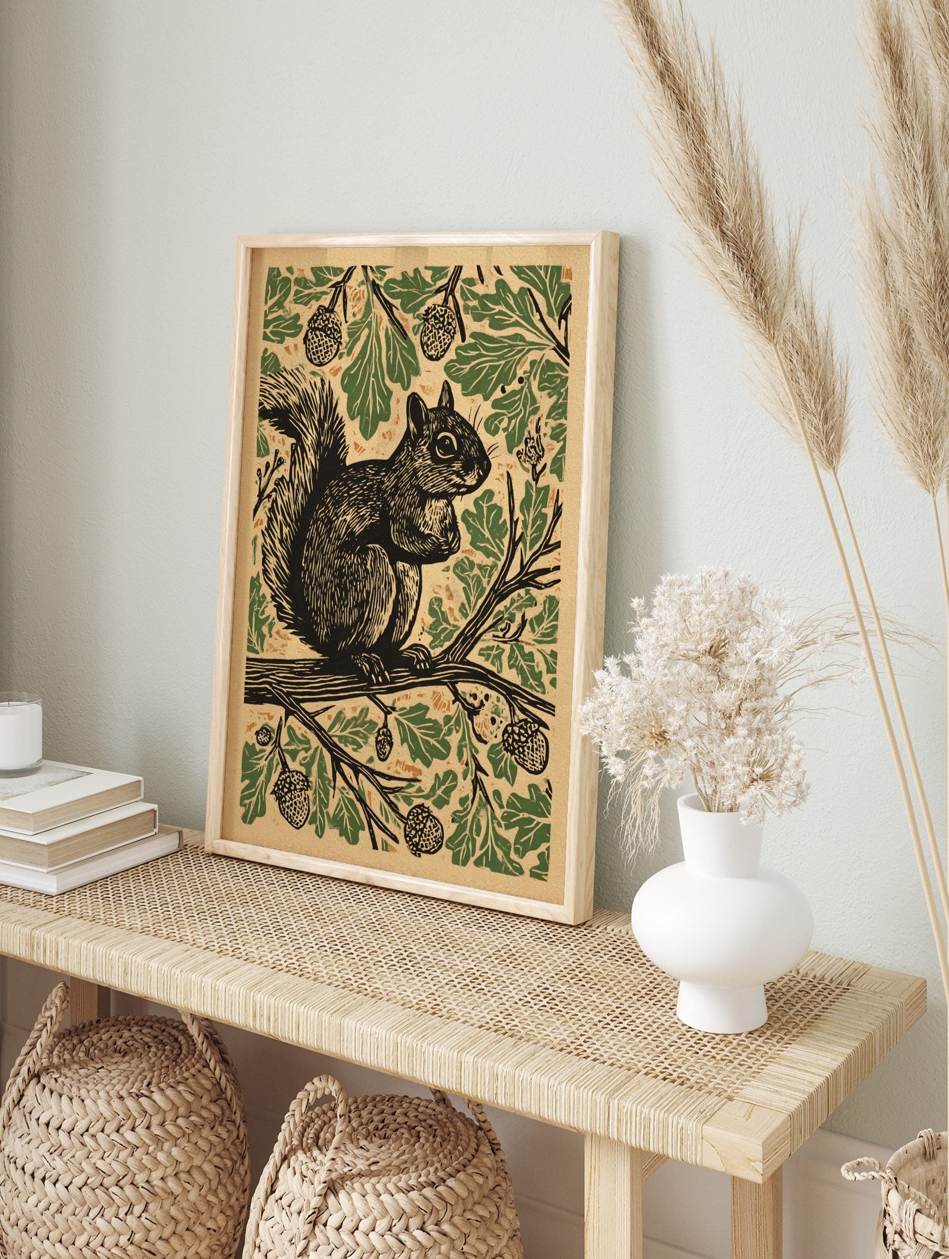 Woodcut style print of a squirrel on an oak branch, surrounded by green leaves and acorns on, in an alternate framed mockup