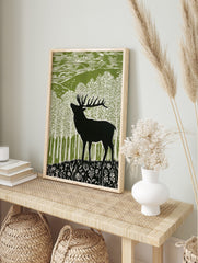 Lino cut style art print of a black stag deer in a green and white forest with hills and, in an alternate framed mockup